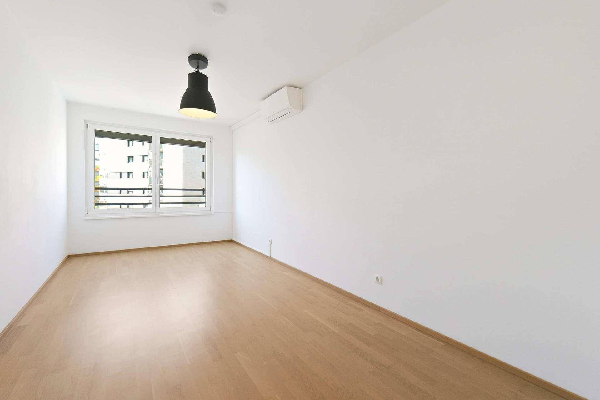 Leeres Schlafzimmer mit hellem Holzboden, großen Fenstern und Klimaanlage.