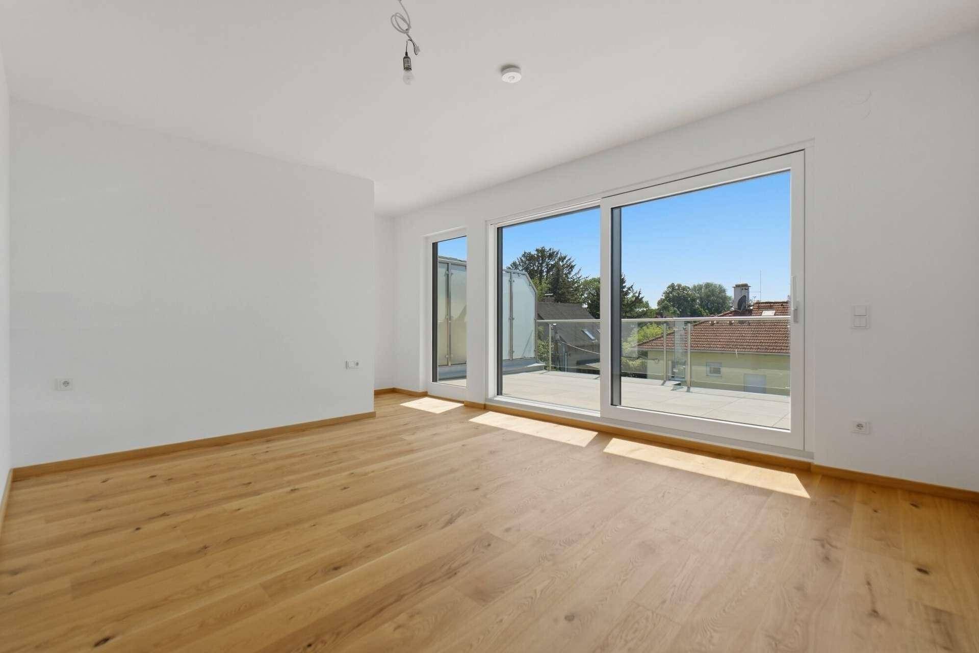 Geräumiges, leeres Zimmer mit Holzboden und großen Fenstern, die auf eine Terrasse führen.