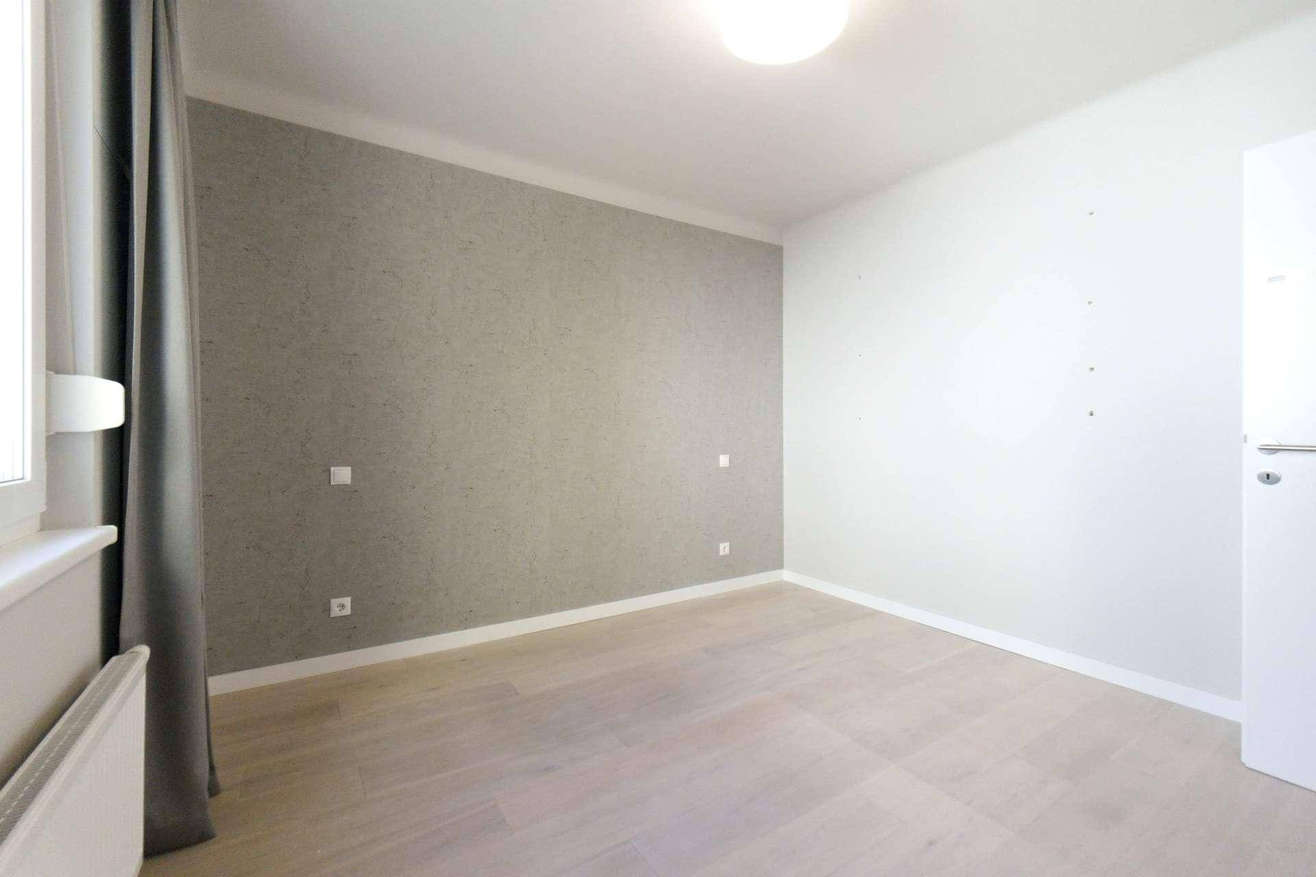 Unmöbliertes Schlafzimmer mit hellem Boden, grauer Akzentwand und Fenster.