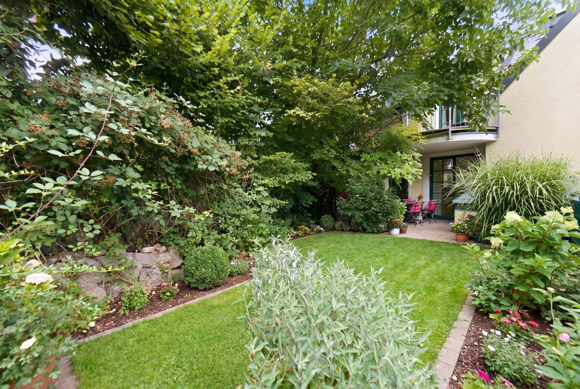 Gepflegter Garten mit Rasenfläche, Sträuchern und einer kleinen Terrasse im Hintergrund.