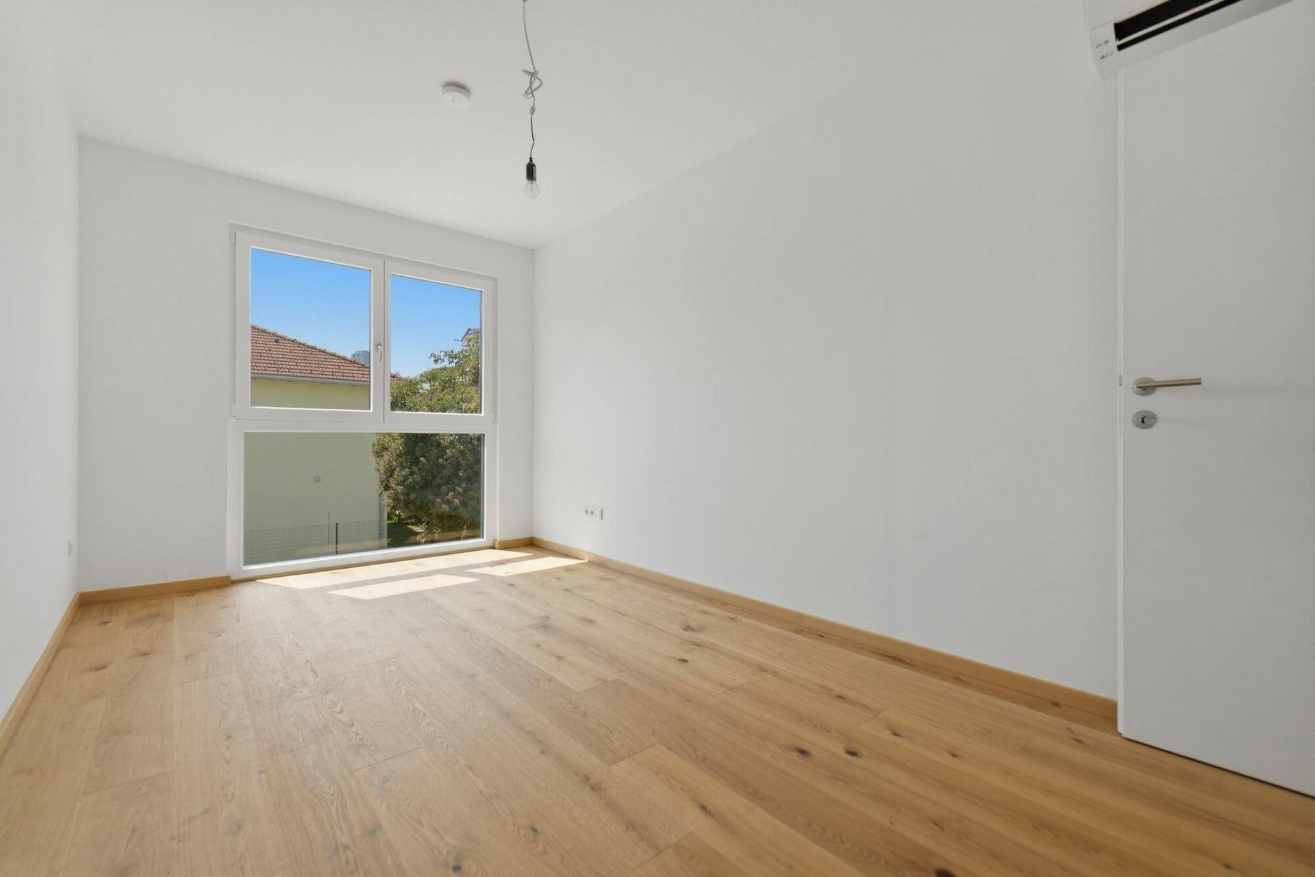 Helles Schlafzimmer mit Holzboden und großem Fenster, das viel Tageslicht hereinlässt.