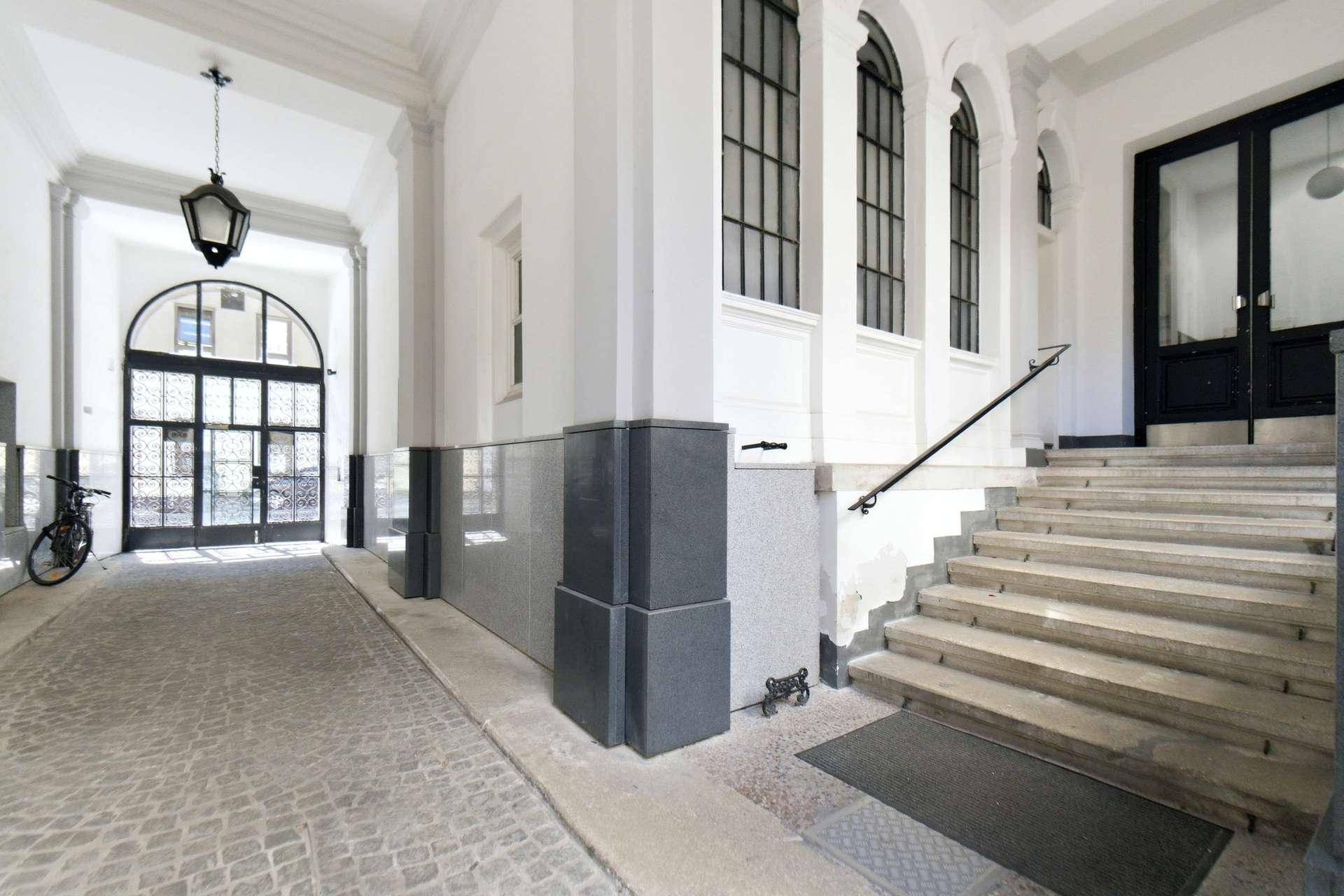 Eingangsbereich des Gebäudes mit gepflastertem Weg, Treppe und großen Bogenfenstern.