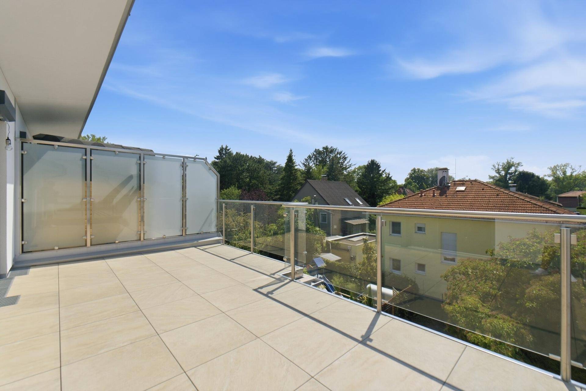 Geräumiger Balkon mit Glasgeländer und weitem Blick über die umliegenden Häuser und Bäume.
