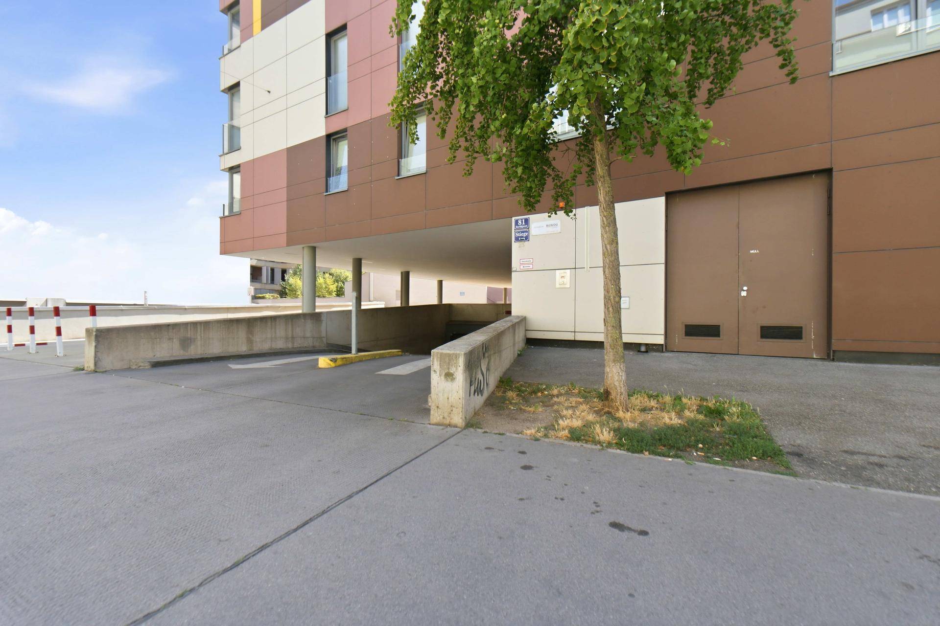 Außenansicht der Tiefgarageneinfahrt eines modernen Wohngebäudes mit Baum und brauner Fassade.