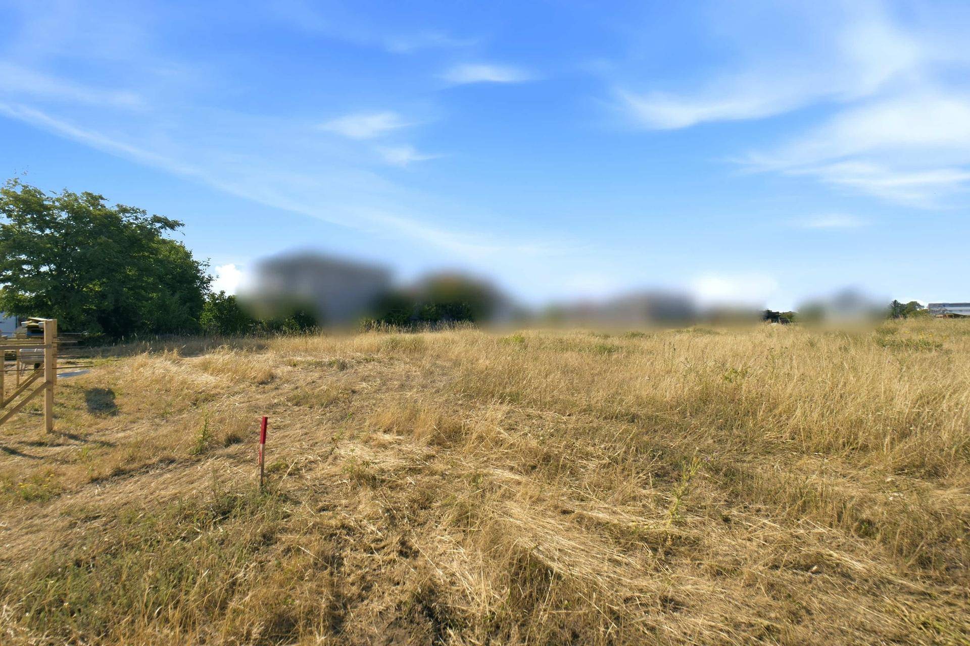 Weites, trockenes Feld unter blauem Himmel mit verschwommenen Gebäuden und einer Holzkonstruktion.