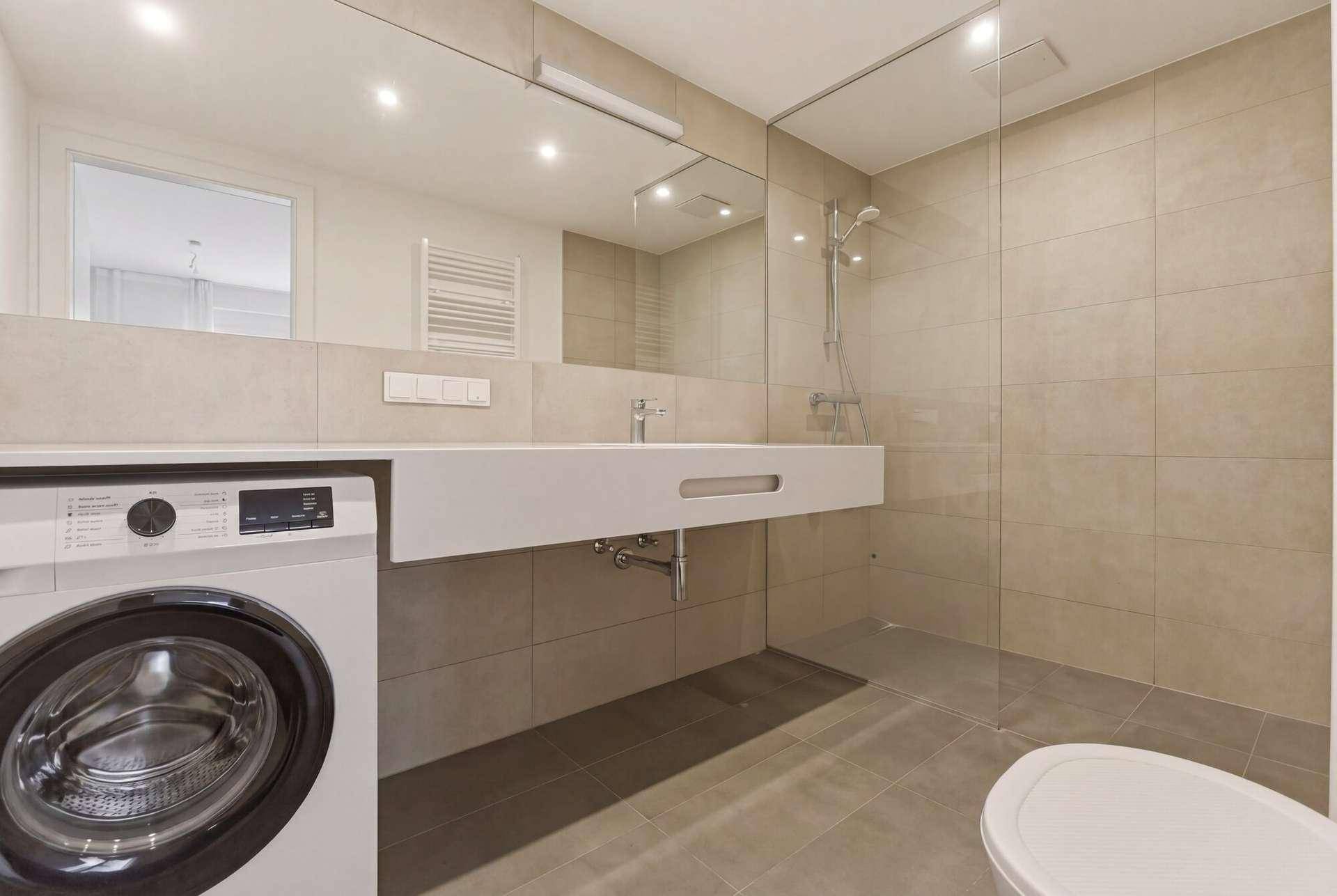 Modernes Badezimmer mit beigefarbenen Fliesen, ebenerdiger Dusche und integrierter Waschmaschine.
