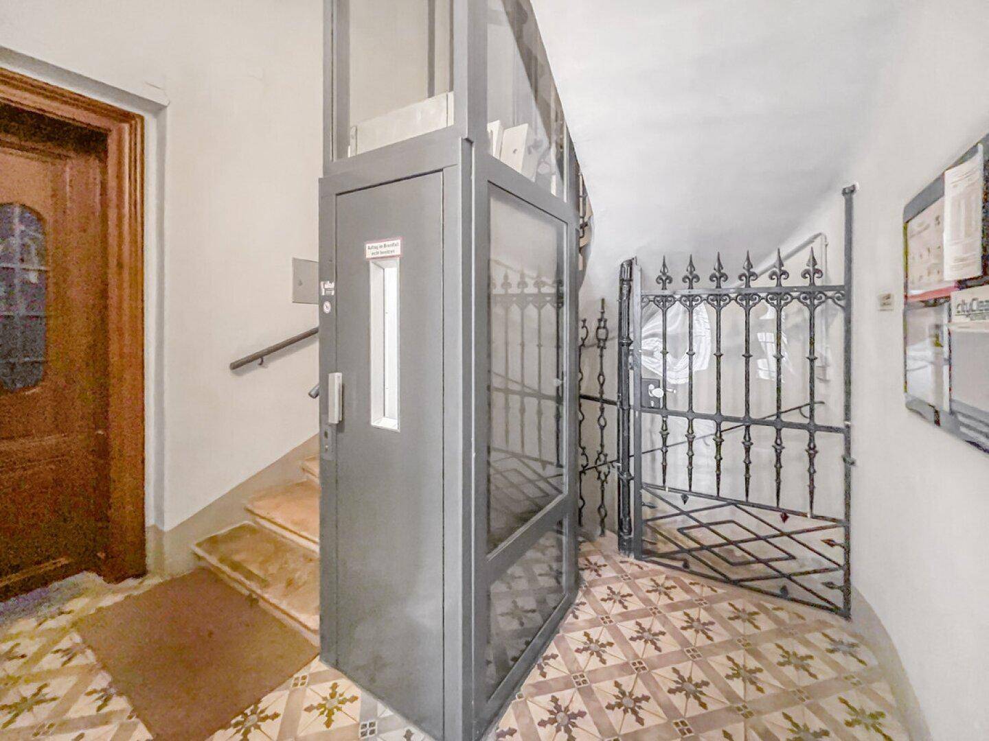 Eingangsbereich mit modernem Aufzug und dekorativen Bodenfliesen in einem Altbau.