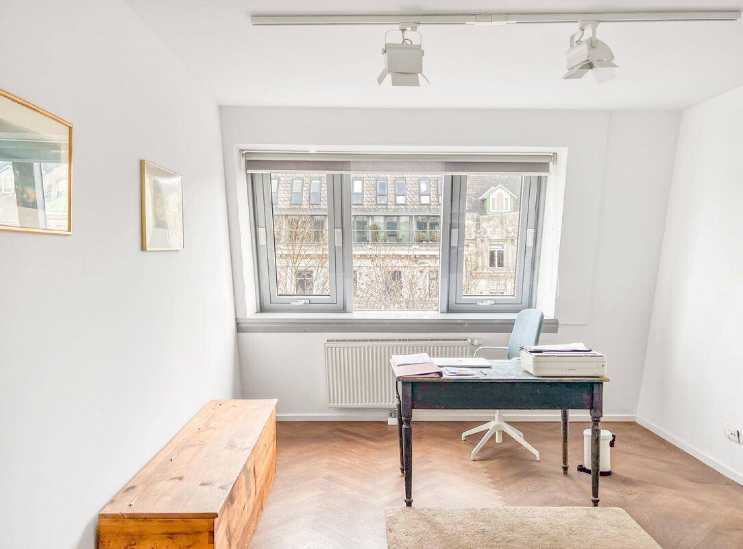Helles Arbeitszimmer mit Schreibtisch, Parkettboden und Blick auf die umliegenden Gebäude.