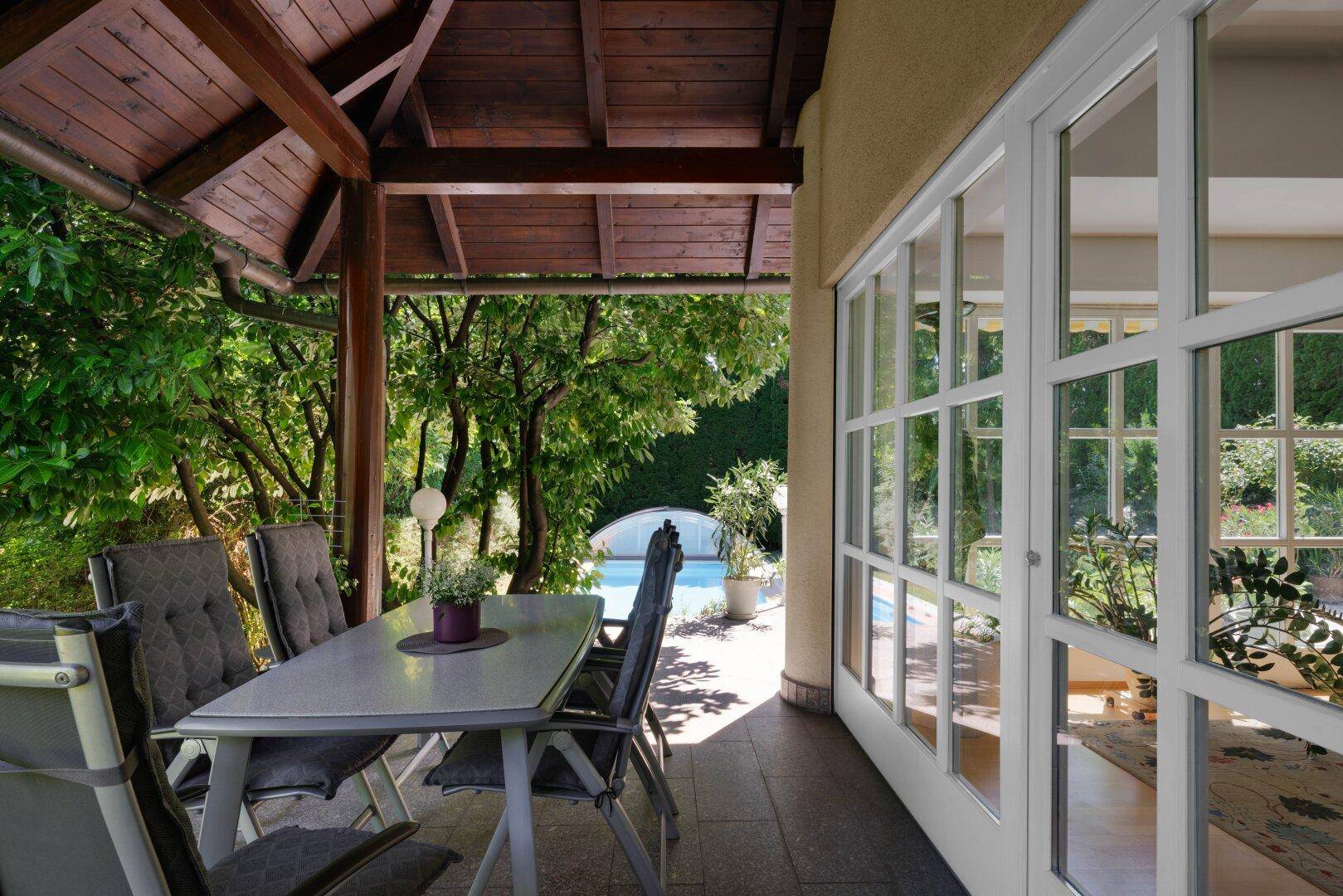 Überdachte Terrasse mit Esstisch und Stühlen, umgeben von üppigem Grün und Poolblick.