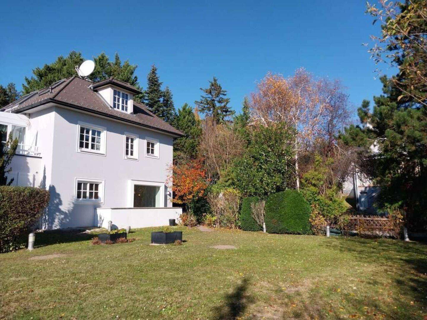 Hausfassade mit großem Gartenbereich, altem Baumbestand und sonnigem Rasen.