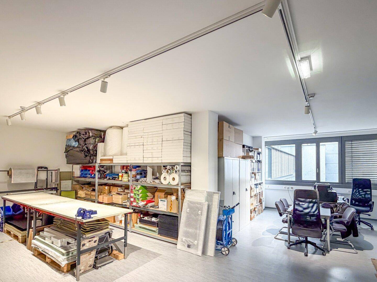 Geräumiger Lagerraum mit Regalen voller Materialien, einem Arbeitstisch und einem angrenzenden Bürobereich.