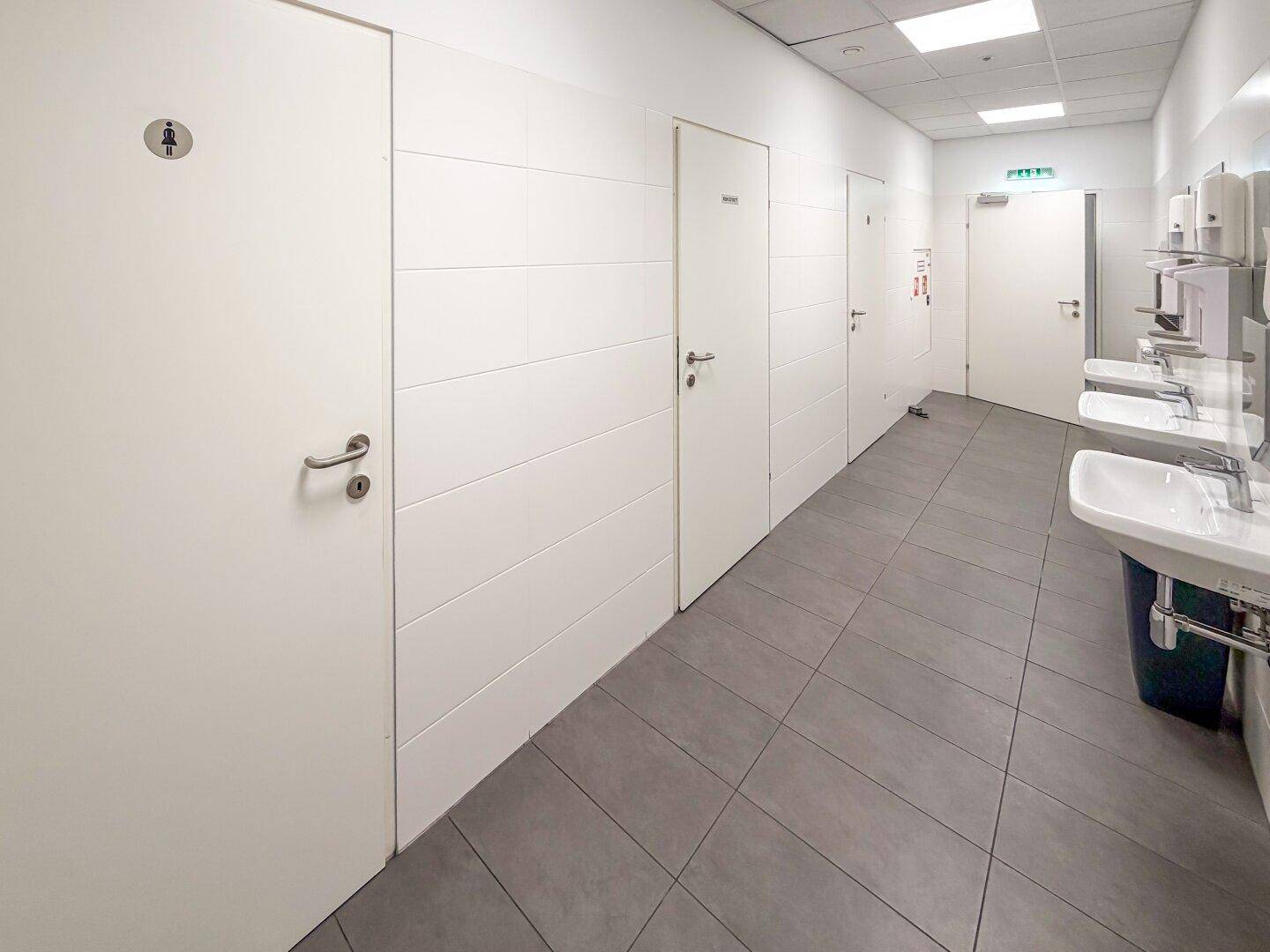 Moderner Waschraum mit mehreren Waschbecken, Spiegeln und getrennten Toilettenkabinen.