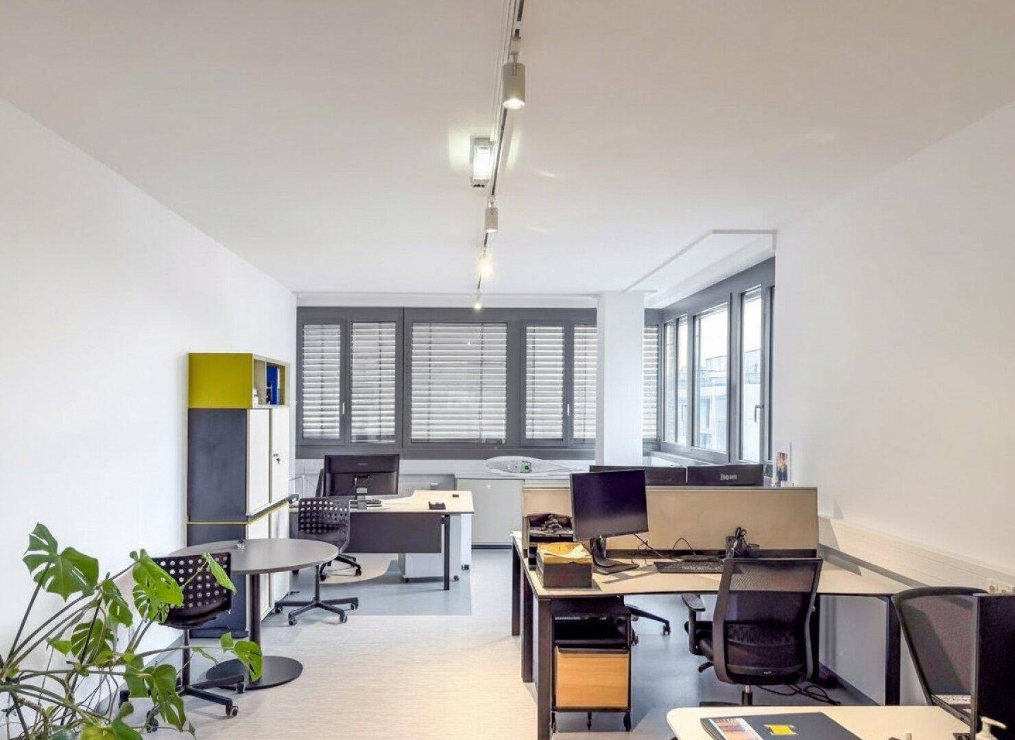 Helles Büro mit mehreren Arbeitsplätzen, modernen Möbeln und einer Pflanze, die für eine angenehme Atmosphäre sorgt.