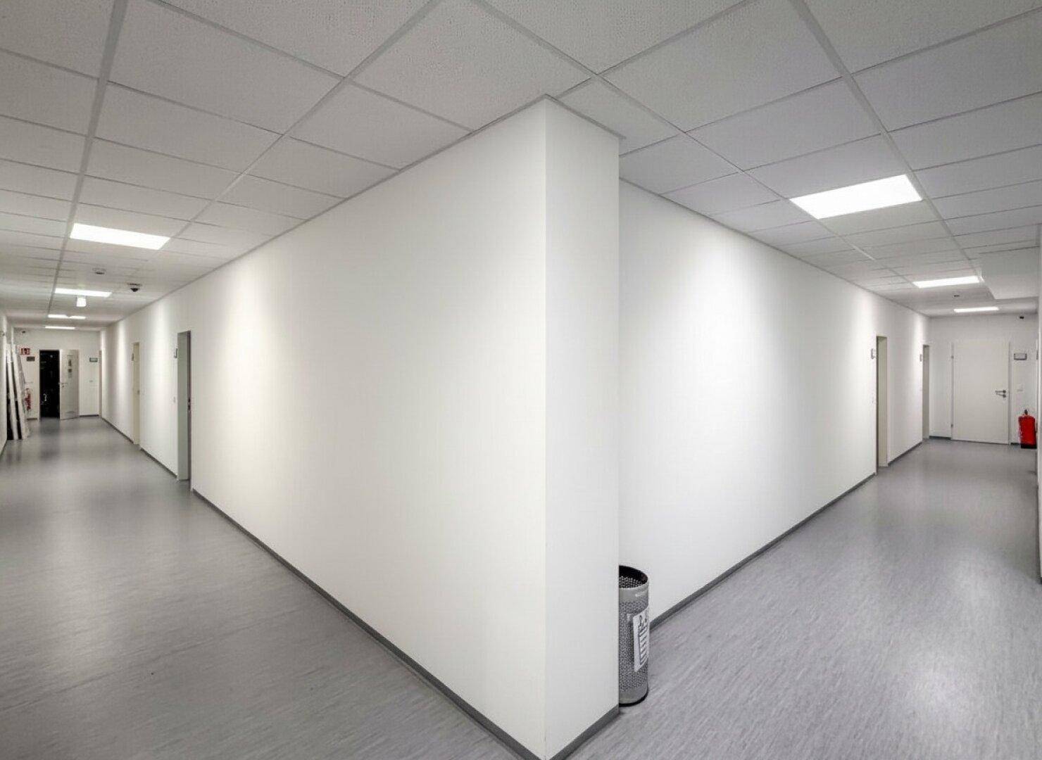 Langer, heller Flur mit weißen Wänden, abgehängter Decke und modernen Deckenleuchten.