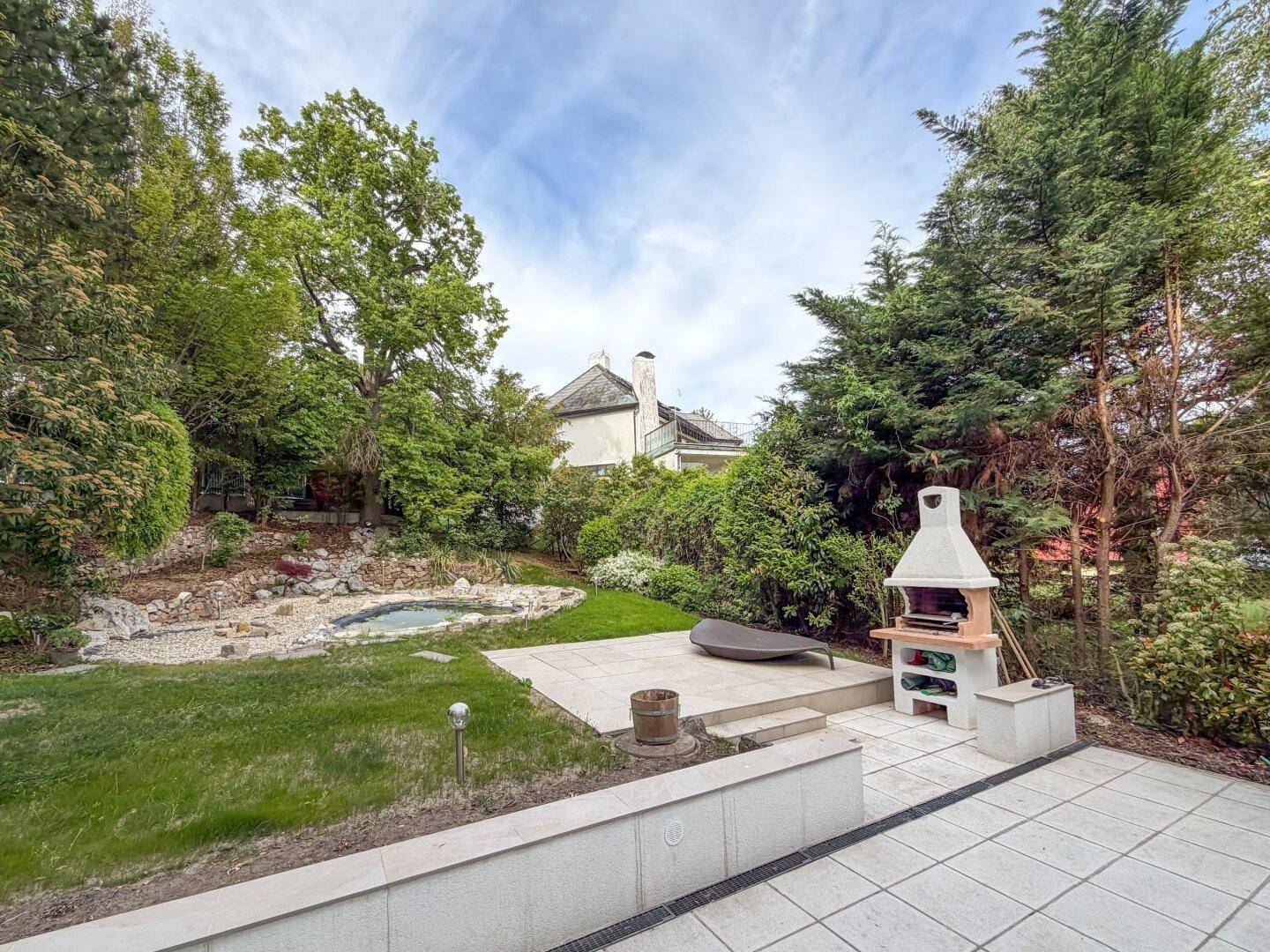 Weitläufiger Gartenbereich mit gepflasterter Terrasse, Grillplatz und Teichanlage.