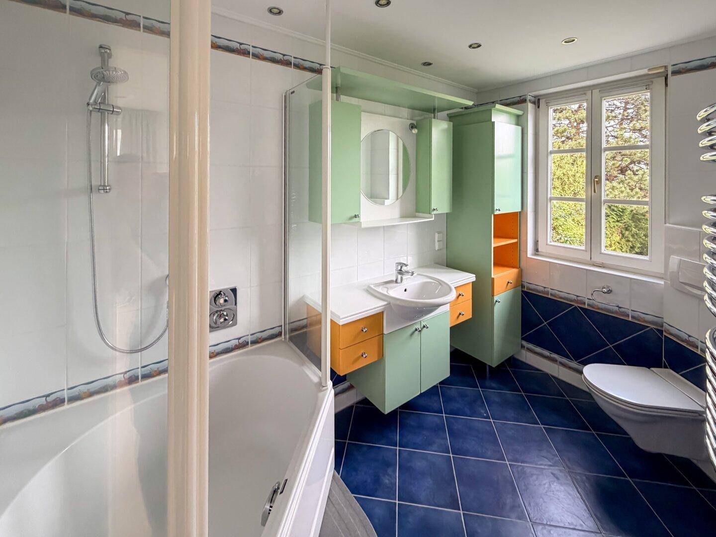 Buntes Badezimmer mit blauen Fliesen, Dusche und Fenster mit Blick ins Grüne.
