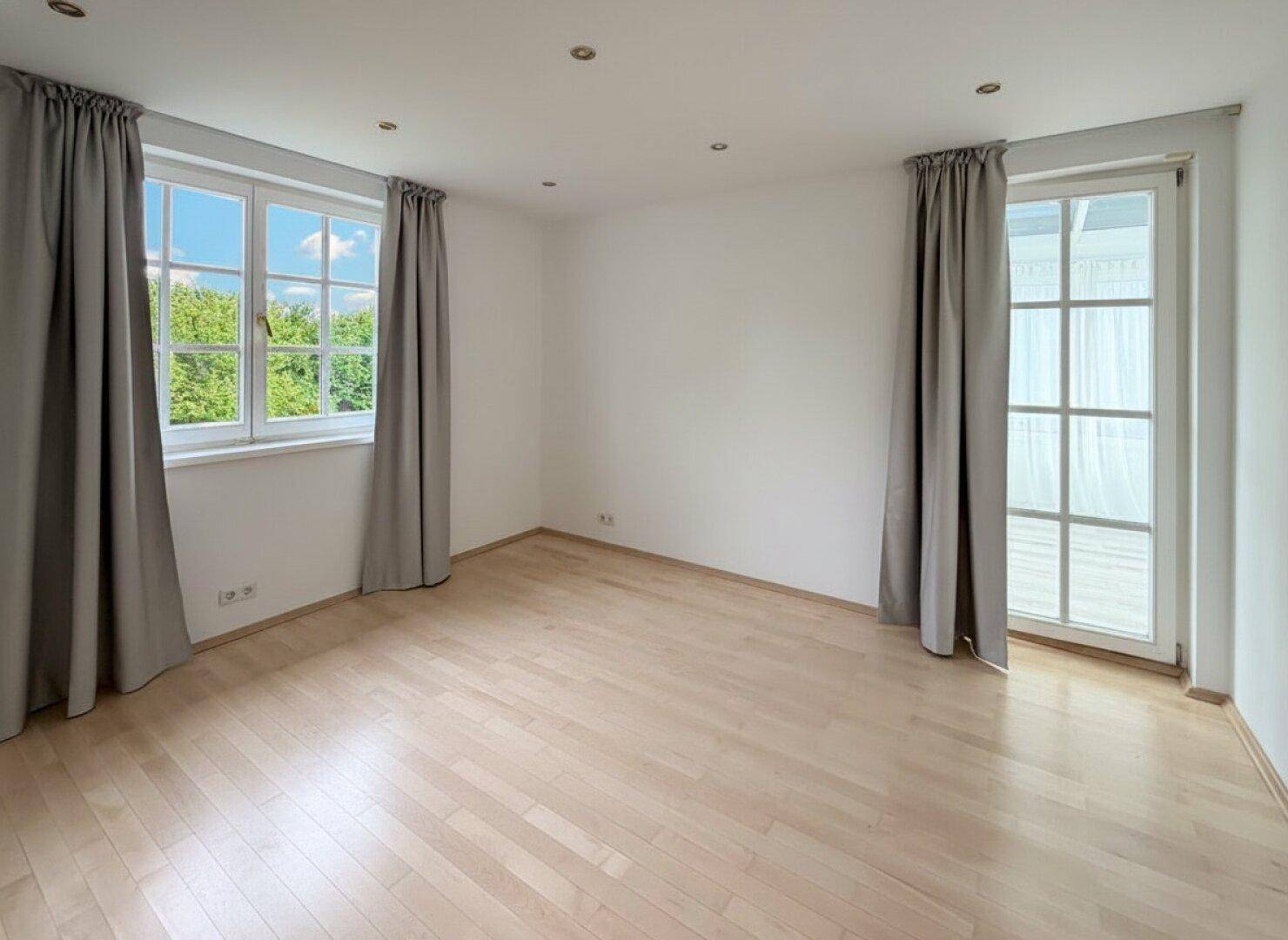 Leerer Raum mit hellem Holzboden, großen Fenstern und viel Tageslicht.
