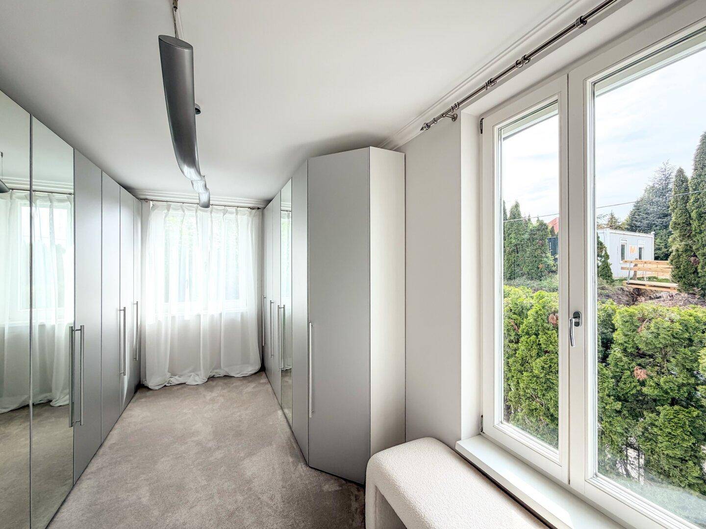Geräumiger begehbarer Kleiderschrank mit Spiegeltüren und Fenster mit Blick ins Grüne.