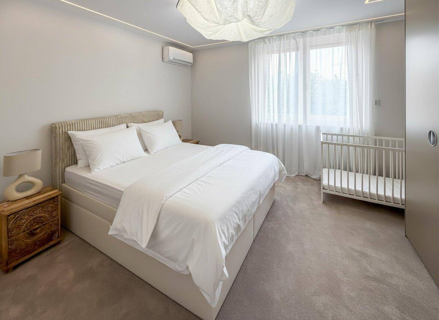 Helles Schlafzimmer mit Doppelbett, Klimaanlage und Babybett am Fenster.