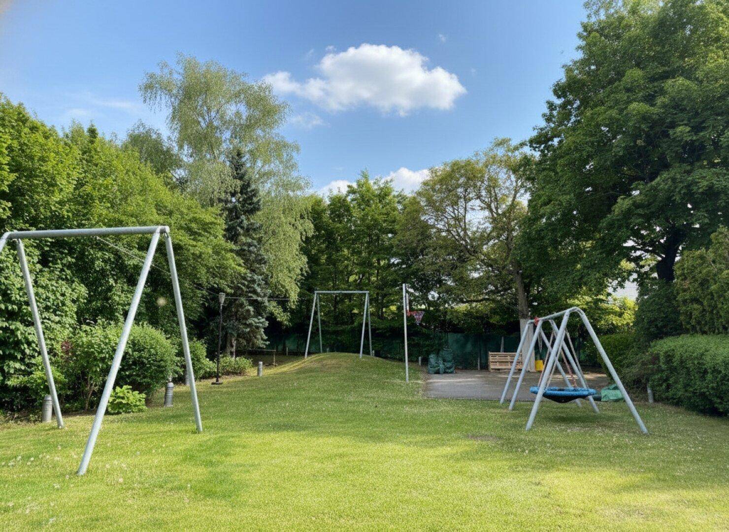 Weitläufiger Garten mit Schaukeln und Spielgeräten für Kinder unter blauem Himmel.