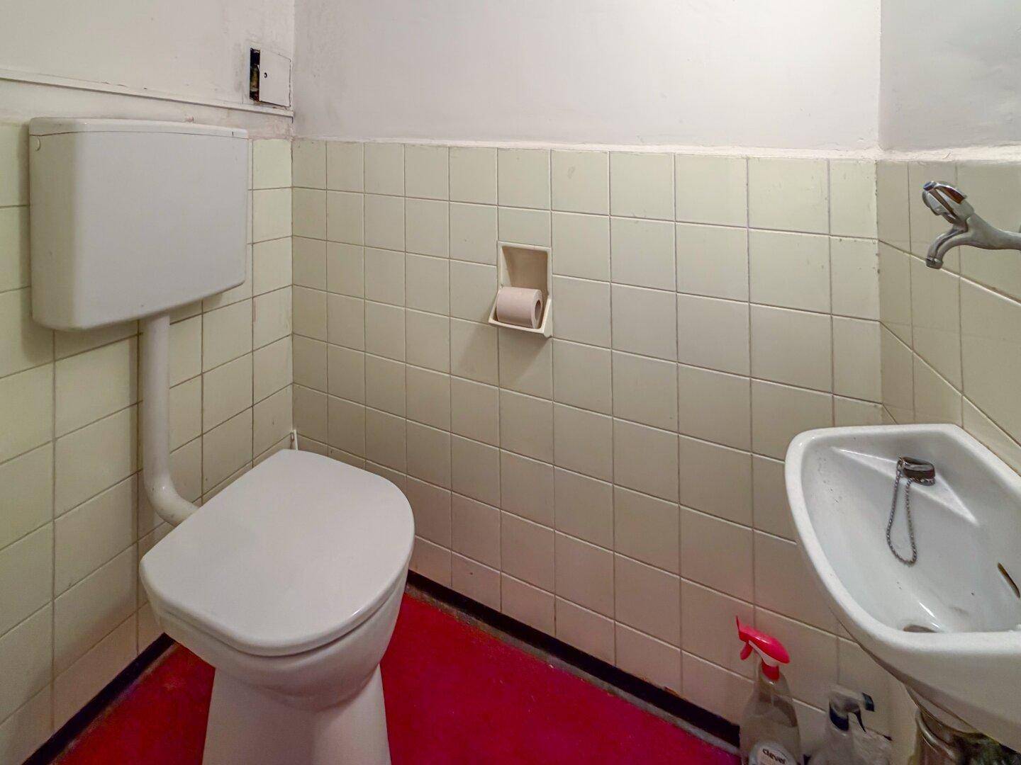 Separates WC mit Toilette und kleinem Waschbecken, ausgestattet mit hellen Fliesen und rotem Boden.