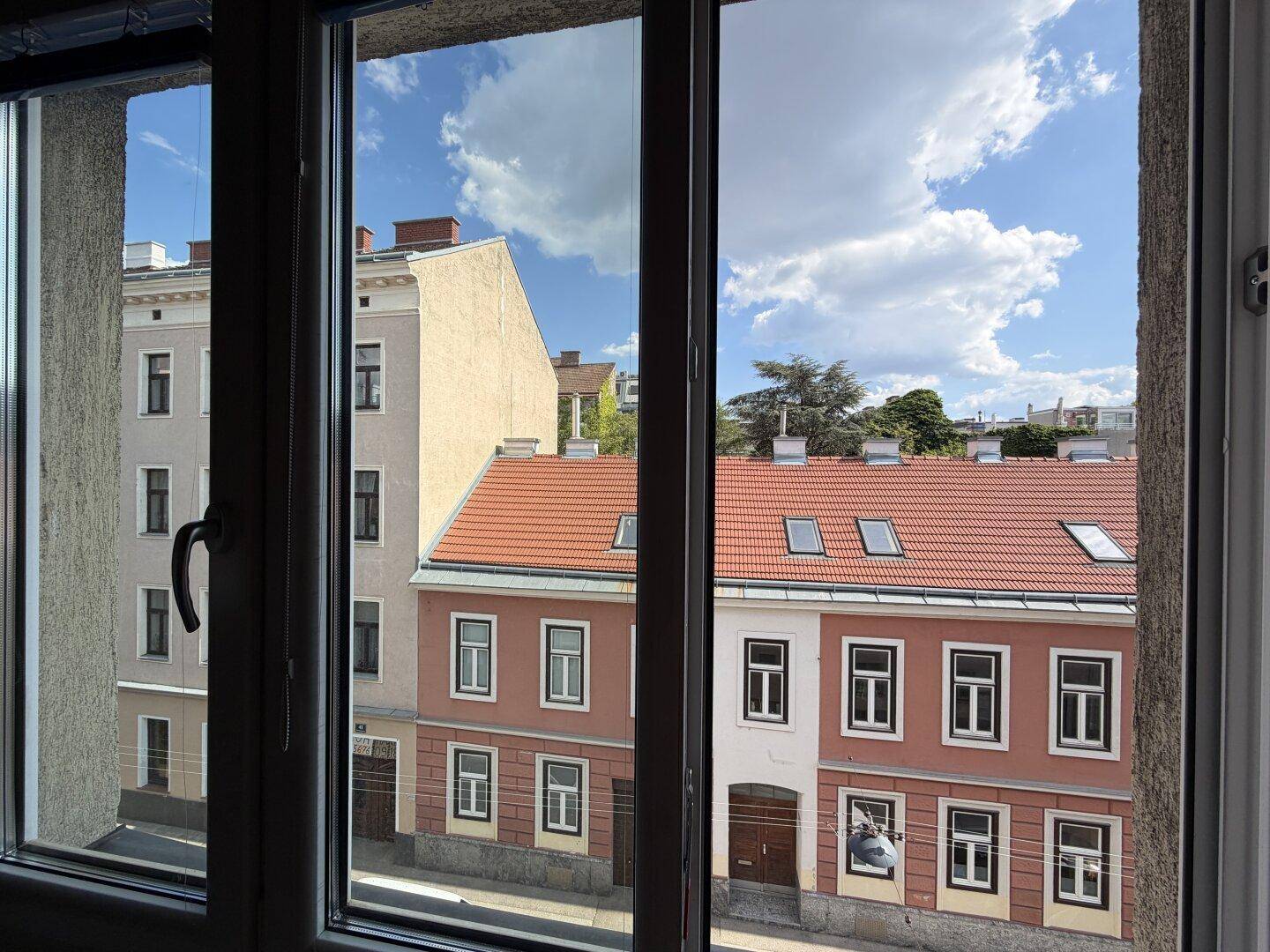 Blick aus einem Fenster auf eine ruhige Straße mit gegenüberliegenden Gebäuden und blauem Himmel.
