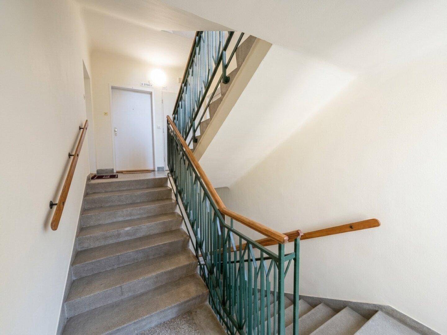 Treppenhaus mit Steinstufen, grünem Metallgeländer und Holzhandlauf in einem Mehrfamilienhaus.