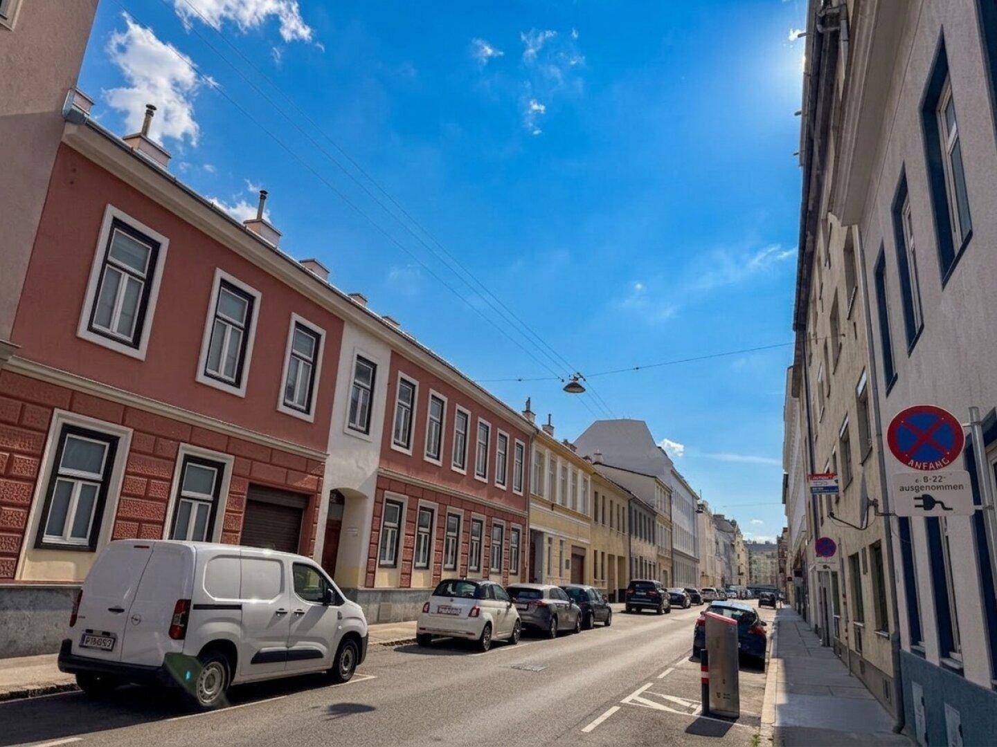 Belebte Wohnstraße mit geparkten Autos und traditionellen Gebäuden unter blauem Himmel.