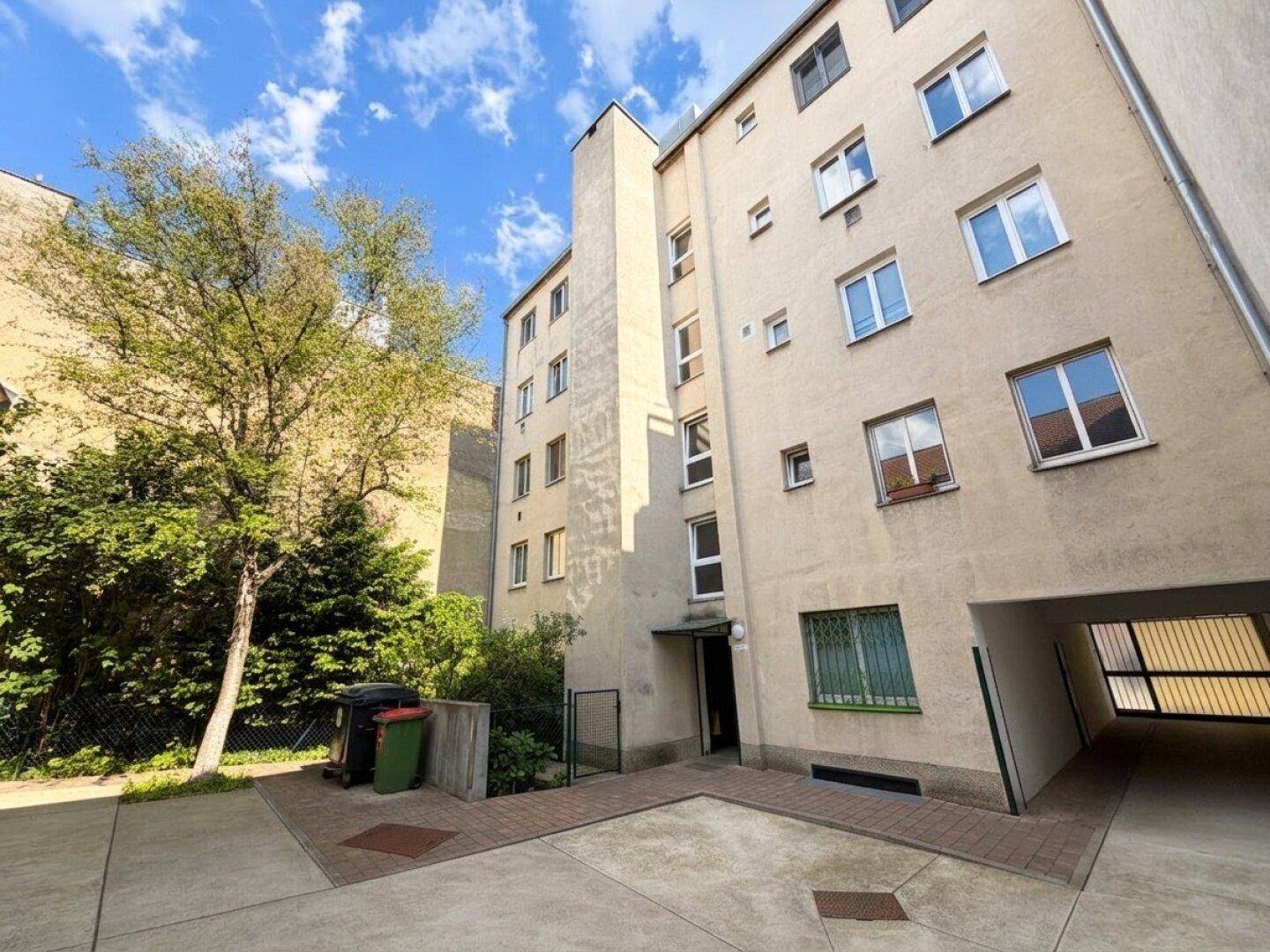 Außenansicht eines Mehrfamilienhauses mit Eingangsbereich, Bäumen und Durchfahrt zum Hof.