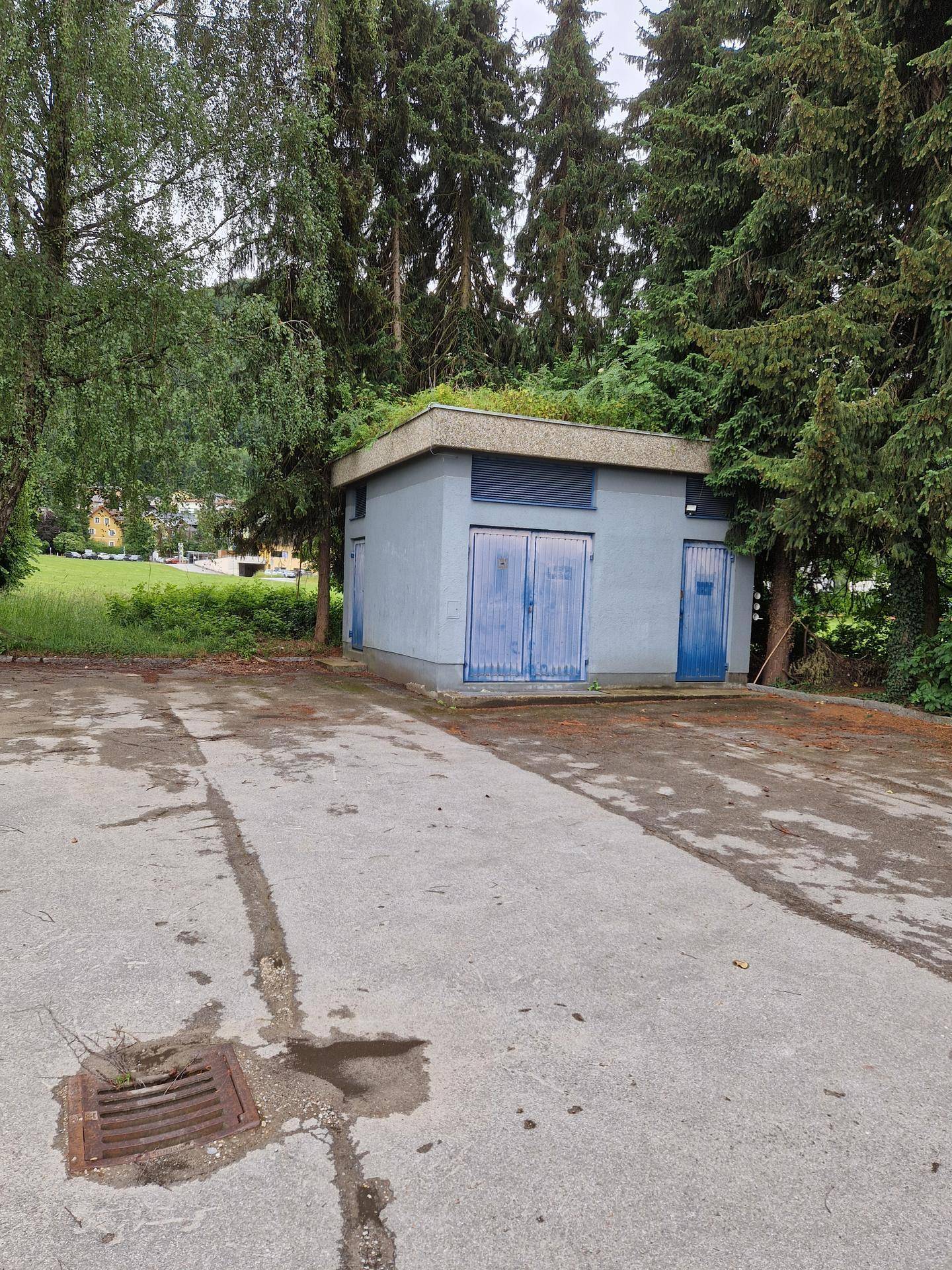 Ansicht eines grauen Funktionsgebäudes mit blauen Türen, umgeben von Bäumen und einem Betonvorplatz.