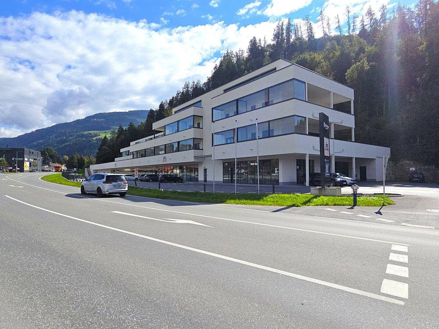Gesamtansicht des modernen Gebäudekomplexes mit Parkplätzen, umgeben von einer malerischen Berglandschaft und viel Grün.