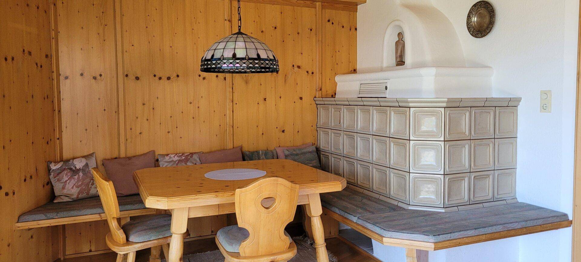 Detailansicht des Essbereichs mit Holztisch, Eckbank und einem traditionellen Kachelofen.