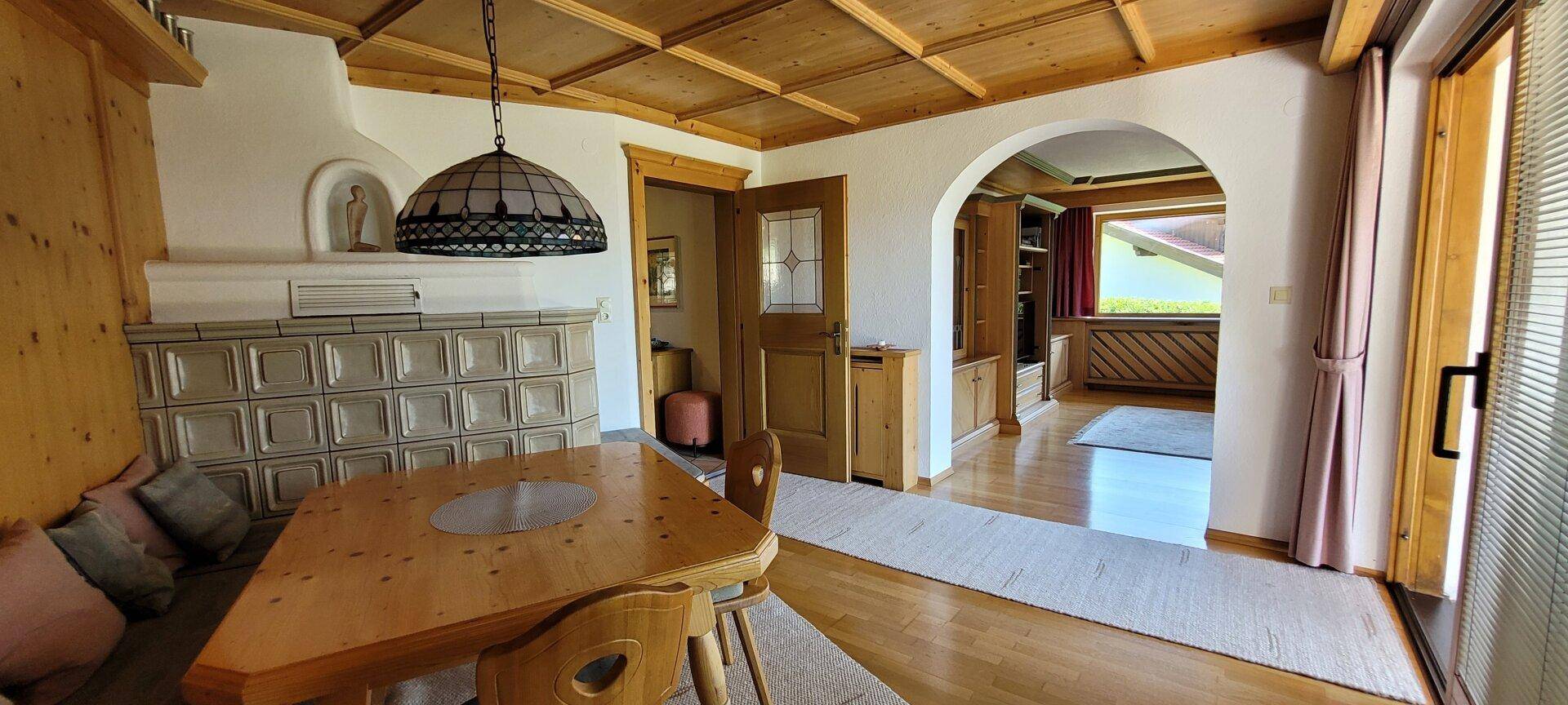 Essbereich mit Holztisch, Eckbank und einem traditionellen Kachelofen, verbunden mit dem Wohnzimmer.