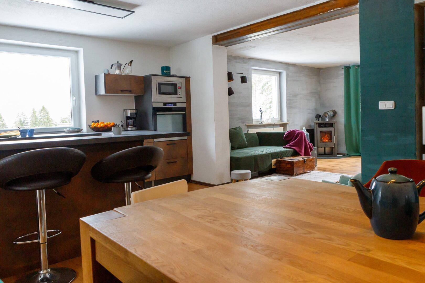 Offener Wohn- und Essbereich mit Holztisch, Kücheninsel und Kaminofen im Hintergrund.