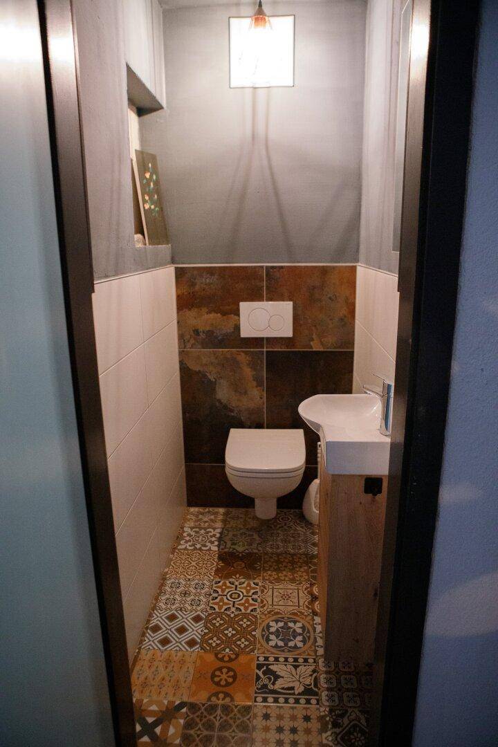 Gäste-WC mit Wand-WC, kleinem Waschbecken und gemusterten Bodenfliesen in Erdtönen.
