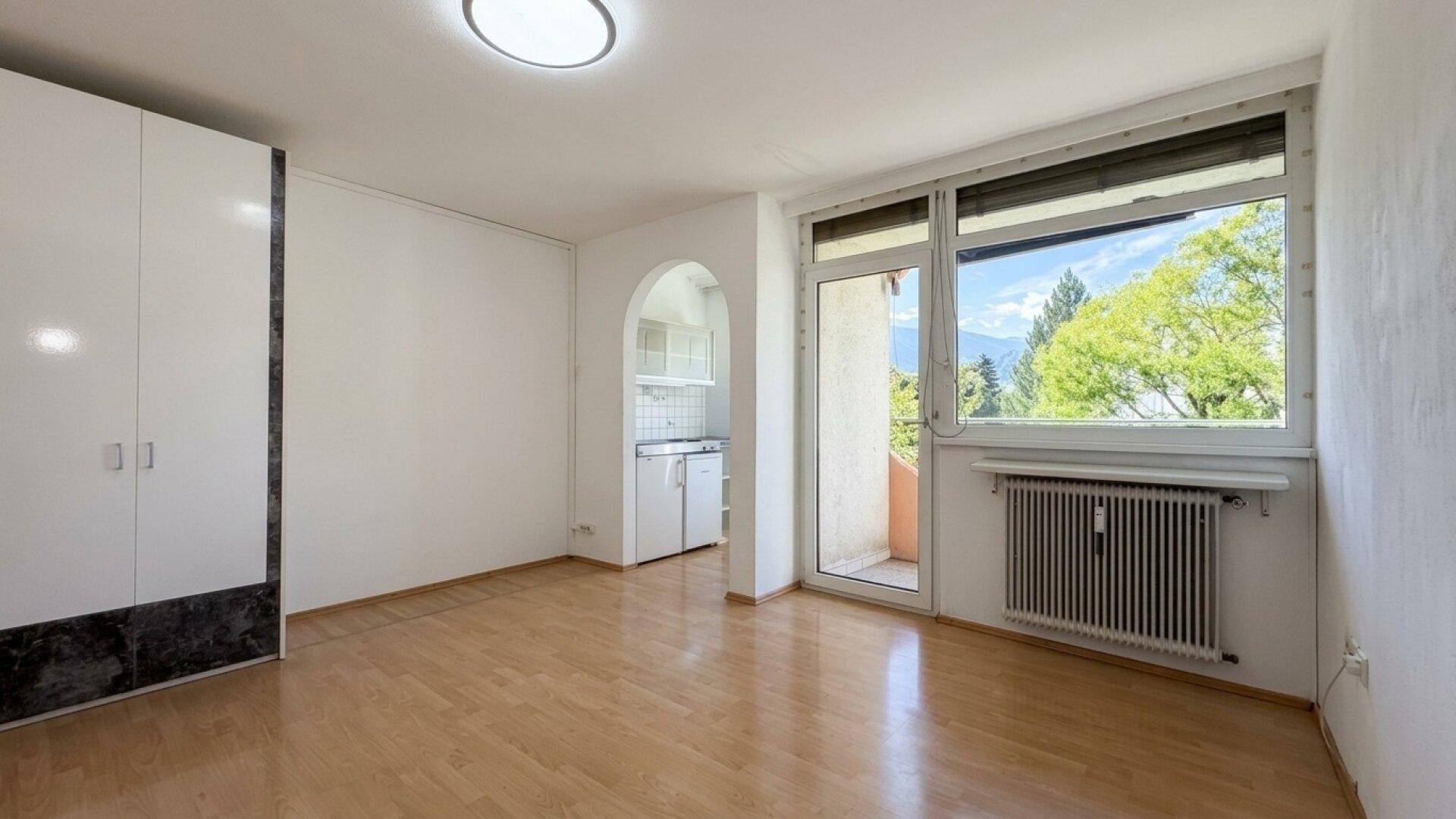 Helles Wohnzimmer mit Parkettboden, großem Fenster und Zugang zu einer Kochnische.