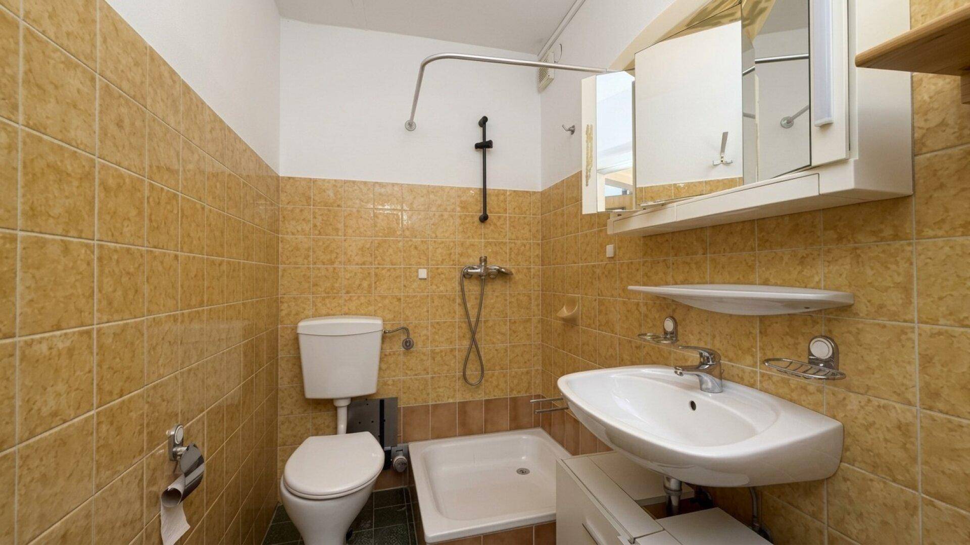 Badezimmer mit gelben Fliesen, Dusche, WC und Waschbecken im 80er-Jahre-Stil.
