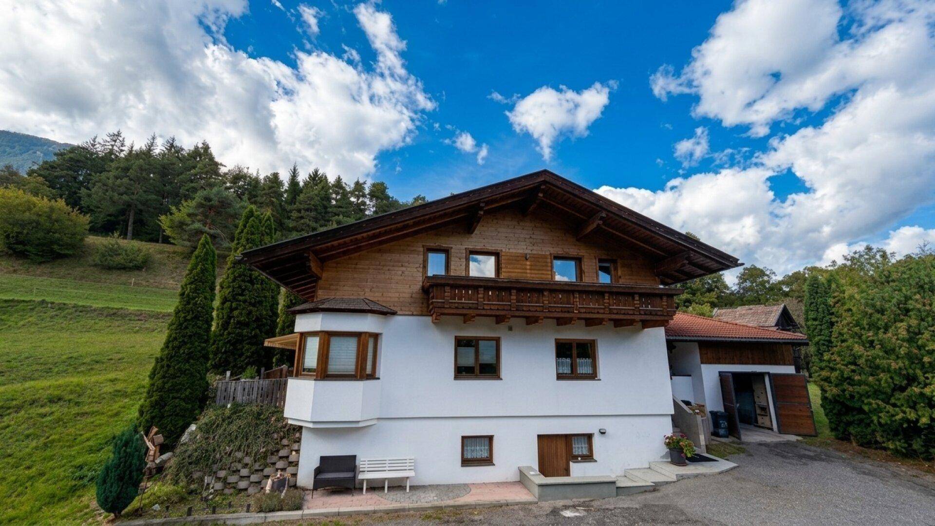 Traditionelles Haus mit weißer Fassade und Holzbalkonen, eingebettet in eine grüne Hügellandschaft.