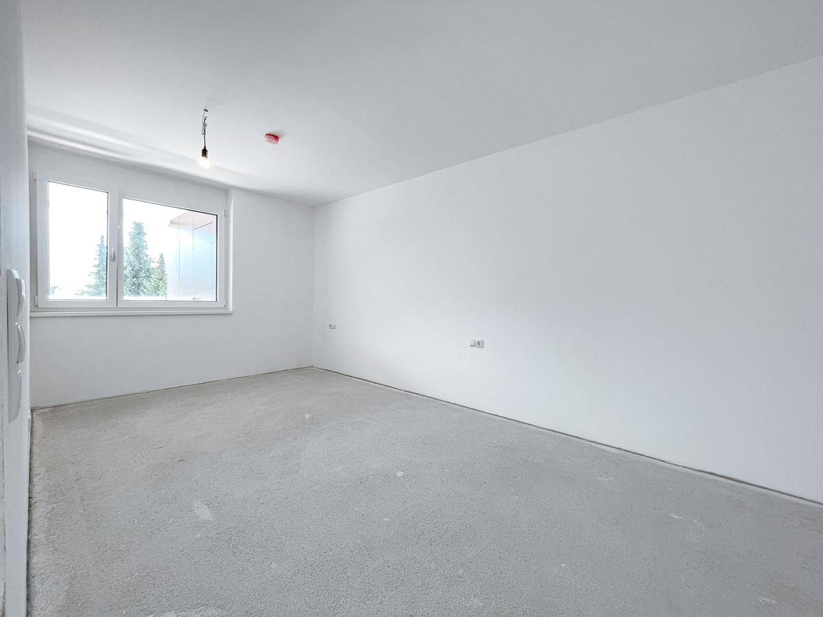Unfertiges Schlafzimmer mit weiß gestrichenen Wänden, Betonboden und einem Fenster für Tageslicht.