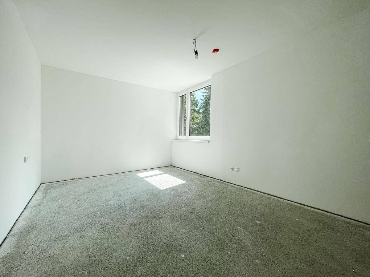 Helles Zimmer im Rohbau mit weiß gestrichenen Wänden, Betonboden und einem Fenster für Tageslicht.