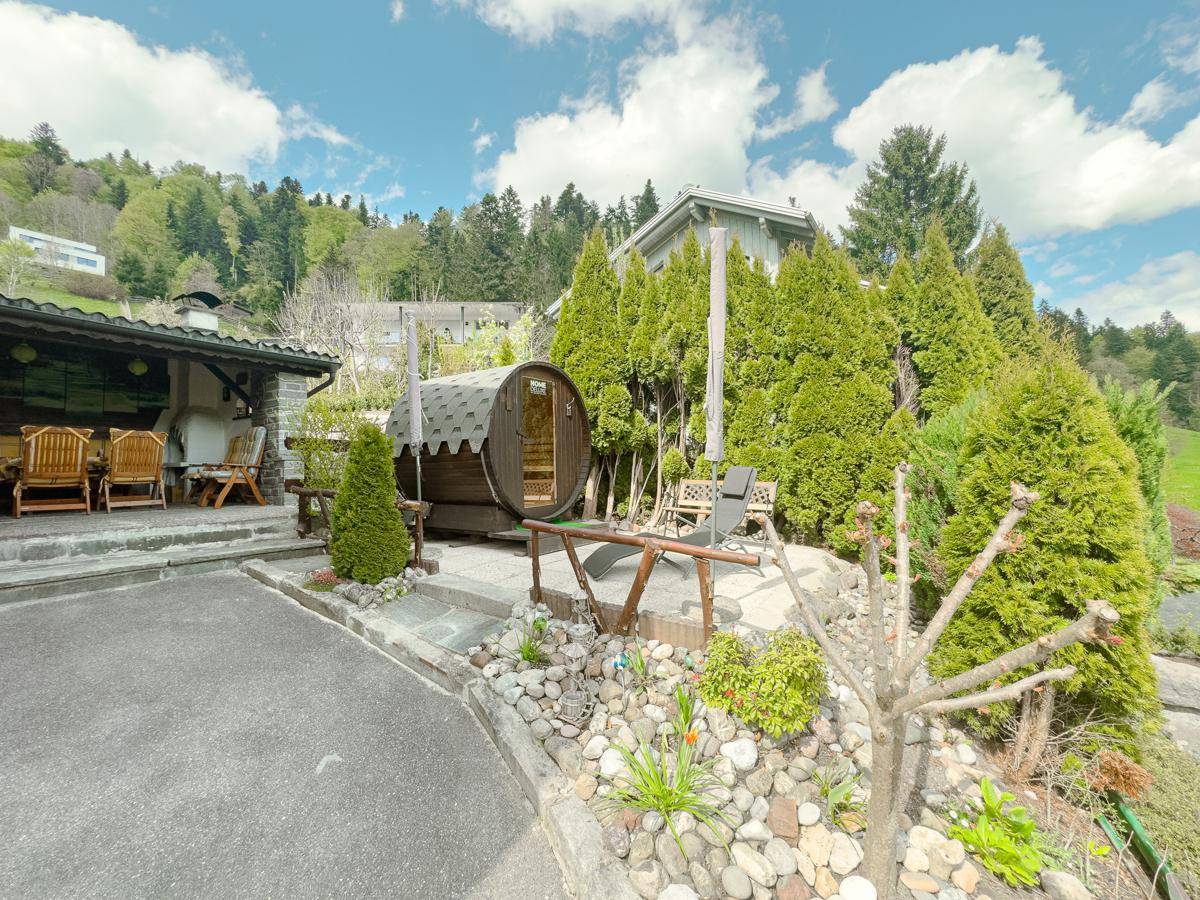 Gartenbereich mit Fasssauna, Steingarten und üppiger Bepflanzung in alpiner Umgebung.