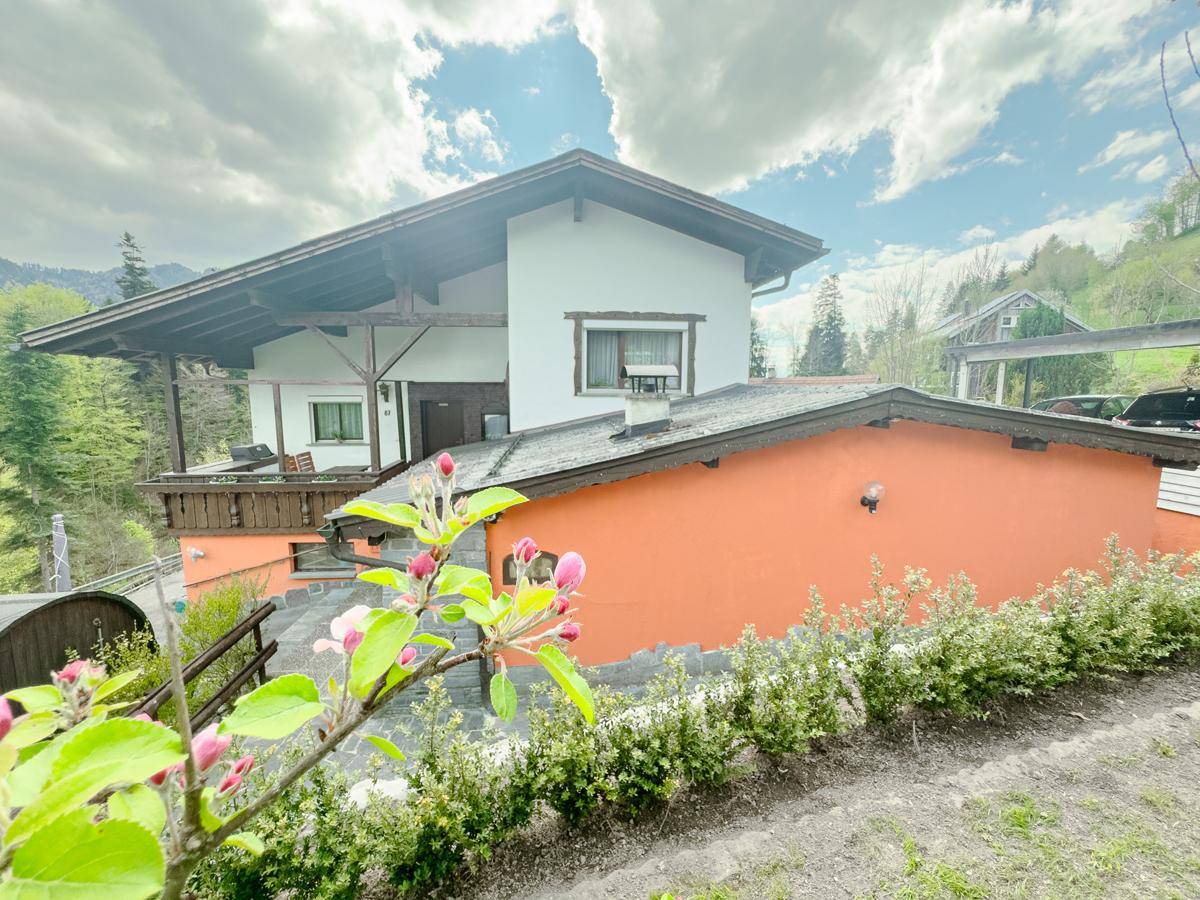 Ansicht des Hauses mit traditionellem Dachüberstand, umgeben von blühenden Bäumen und Natur.