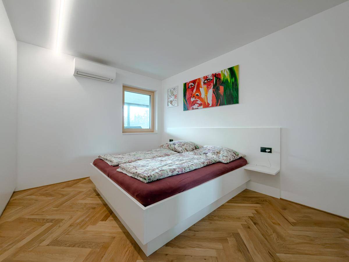 Schlafzimmer mit hellem Doppelbett, Fischgrätparkett, Klimaanlage und dekorativem Wandbild.