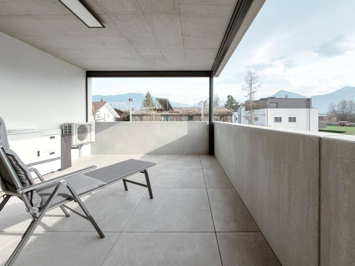 Überdachte Terrasse mit Liegestuhl, Außenmöbeln und Blick auf die umliegende Wohngegend und Berge.