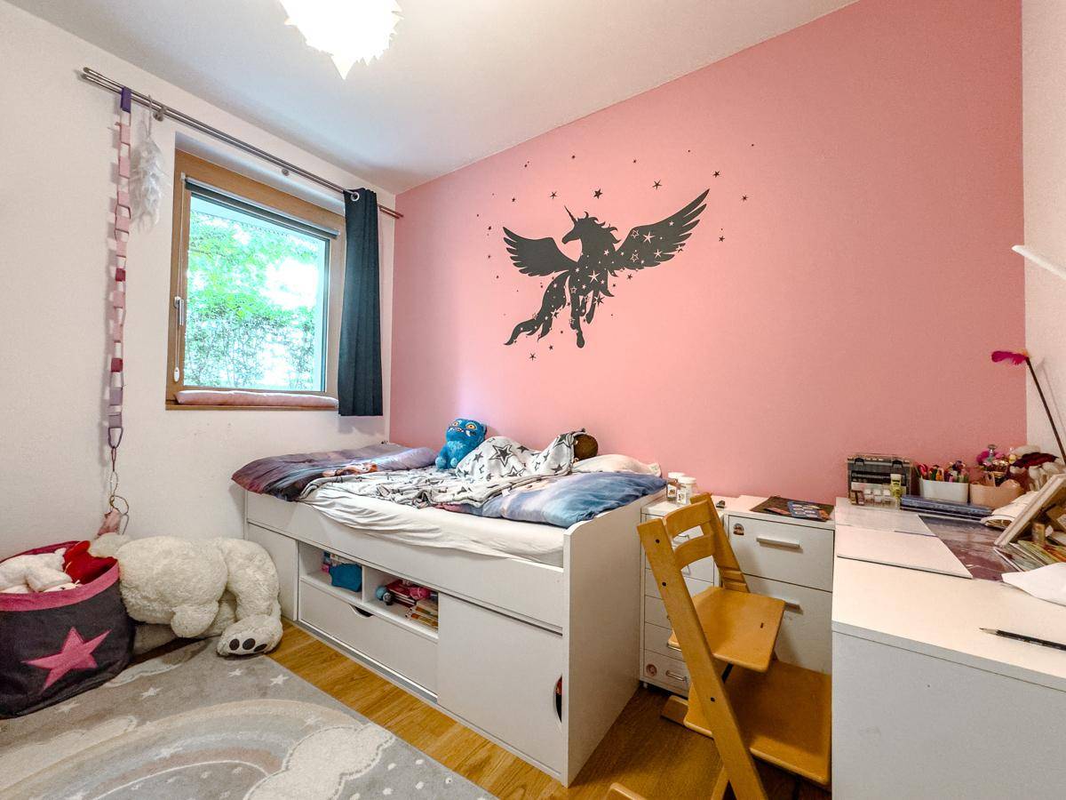 Helles Kinderzimmer mit rosa Wand, Einzelbett und Schreibtisch, mit Blick ins Grüne.
