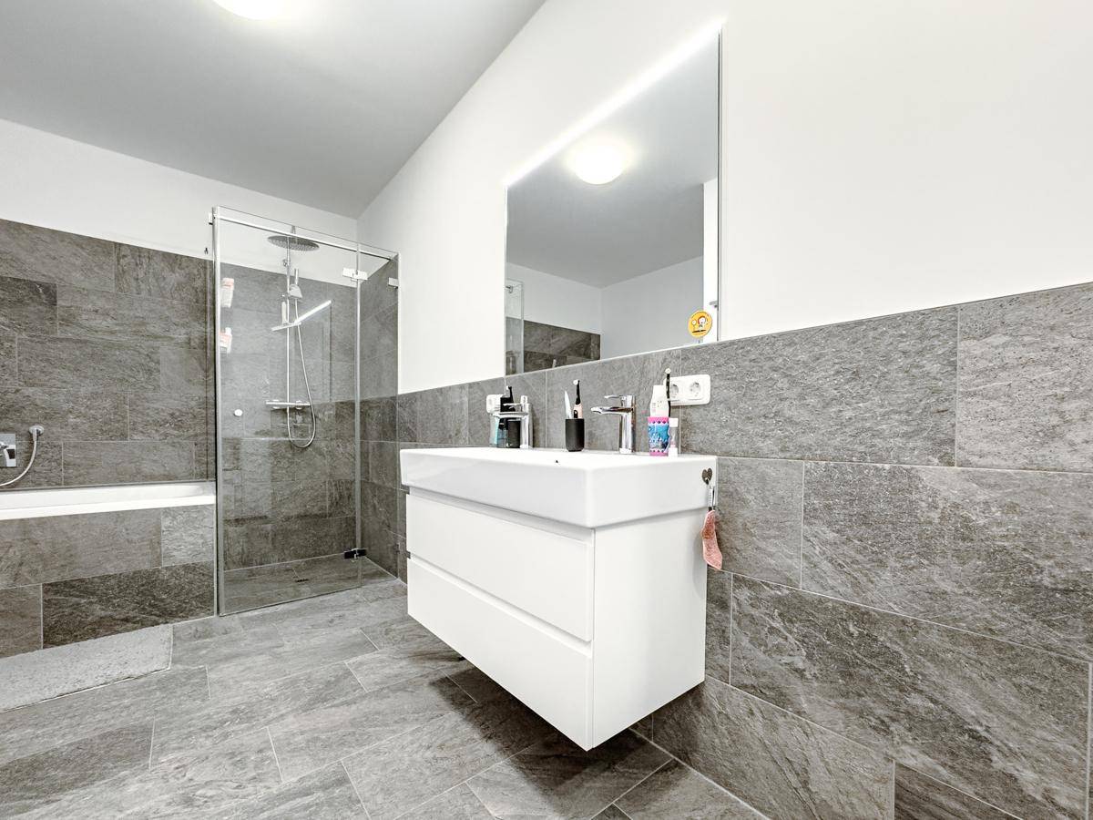 Modernes Badezimmer mit grauen Fliesen, schwebendem Waschtisch und begehbarer Dusche, hell und funktional.