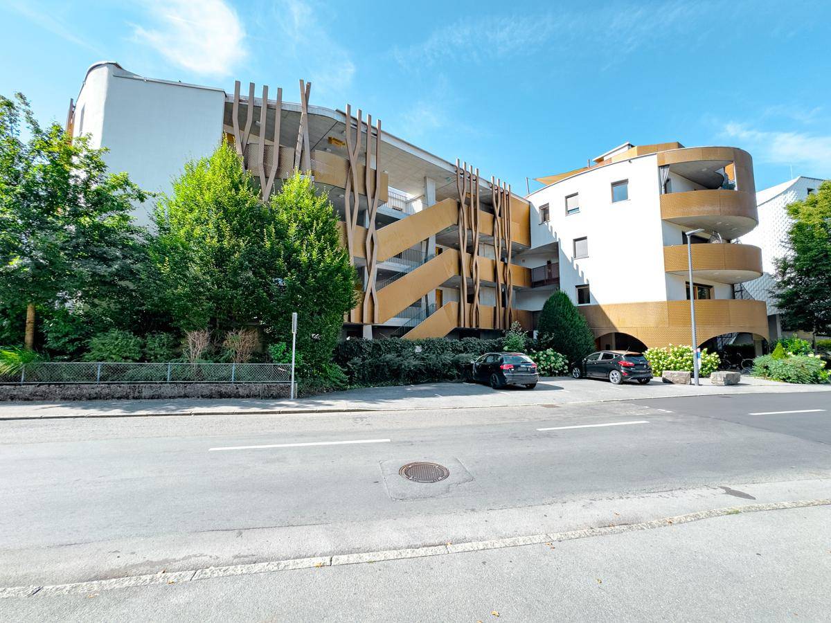 Ansicht des modernen Wohngebäudes von der Straße mit Parkplätzen und viel Grün, unter blauem Himmel.