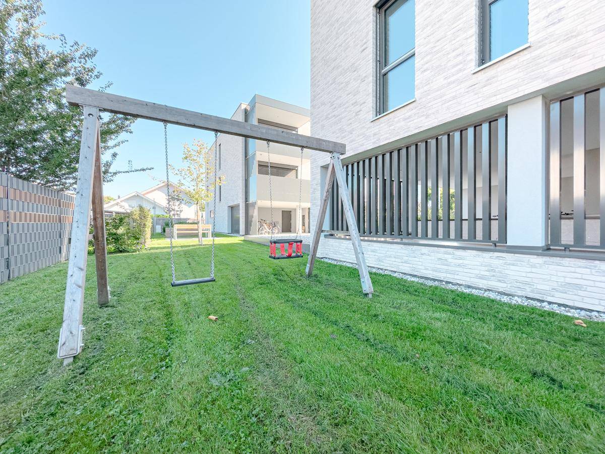 Großer Gartenbereich mit Schaukel und Rasenfläche, umgeben von einem modernen Wohngebäude.