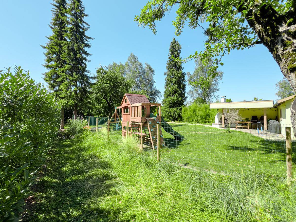 Weitläufiger Garten mit Kinderspielplatz, hohen Bäumen und natürlicher Bepflanzung unter blauem Himmel.