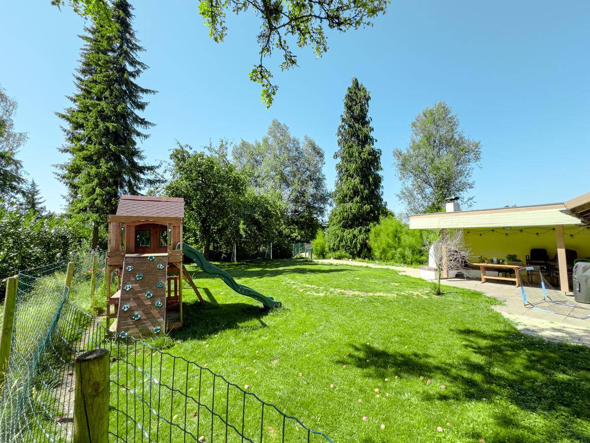 Sonnige Gartenfläche mit Kinderspielplatz, hohen Bäumen und einem überdachten Bereich für Mahlzeiten im Freien.
